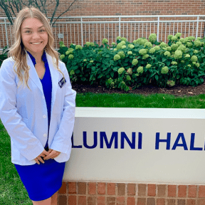Student posing in white coat outside academic building sign