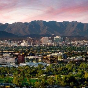 Downtown Phoenix skyline with surrounding greenery and mountains in the background under a pink and blue sunset sky.