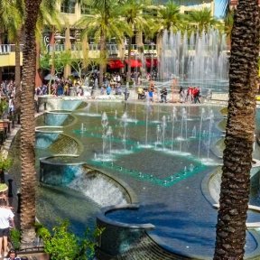 Crowds gather around a large tiered fountain surrounded by palm trees, shops, and restaurants at an outdoor plaza on a sunny day.