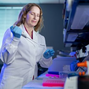 A healthcare professional in a lab setting uses a large dropper to deposit liquid into a test tube.