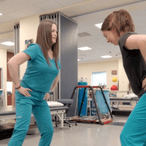 A physical therapist in teal scrubs guides a patient through a bending exercise in a rehabilitation gym.