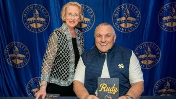 Dr. Kathleen Goeppinger and Rudy Ruetttiger smiling. Rudy holds his book titled "Rudy."