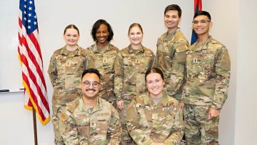 A group of military students smile in their uniforms next to the US flag
