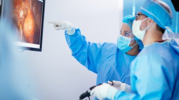 Two surgeons in full surgical gowns, masks, and gloves review a laparoscopic image on a monitor during a procedure.