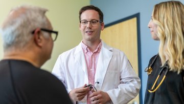 A physician in a white coat holds a stethoscope while speaking with a patient and a medical student in a clinic exam room.