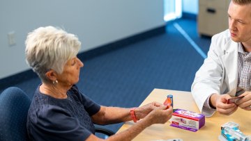 A pharmacy student in a white coat reviews medications with an elderly female patient at a table during a consultation.