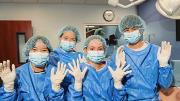 Four students in blue surgical gowns, masks, hair caps, and gloves pose together in a clinical training room.