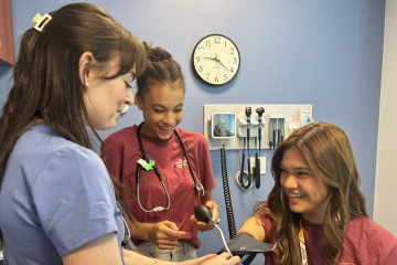 Two high school students learn how to take vital signs from a Midwestern student in the simulation exam room 