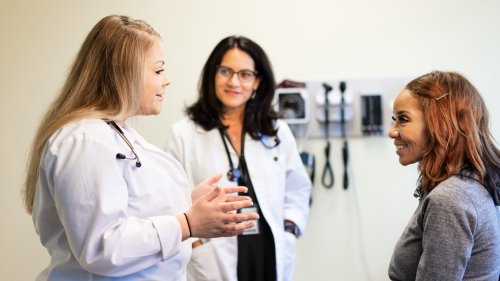 A medical student and supervising physician in white coats speak with a smiling patient in a clinical exam room.