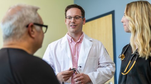 A physician in a white coat holds a stethoscope while speaking with a patient and a medical student in a clinic exam room.