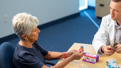 A pharmacy student in a white coat reviews medications with an elderly female patient at a table during a consultation.