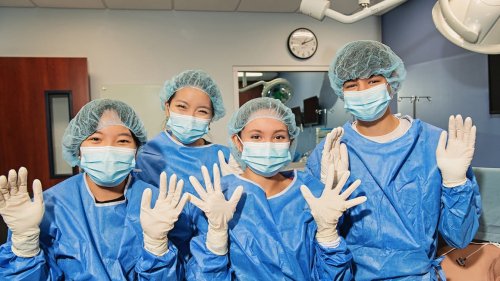 Four students in blue surgical gowns, masks, hair caps, and gloves pose together in a clinical training room.