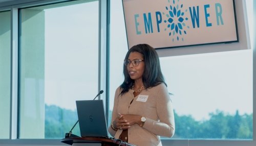 Woman speaking at a podium with a laptop, in front of a window and a screen displaying the word "EMPOWER."