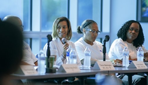 Woman speaking into a microphone during a panel discussion, seated with others at a table with name cards, water bottles, and microphones.