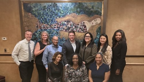 Group of eleven professionally dressed people smiling in front of a colorful painting at an event.