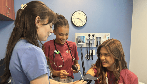 Two high school students learn how to take vital signs from a Midwestern student in the simulation exam room 