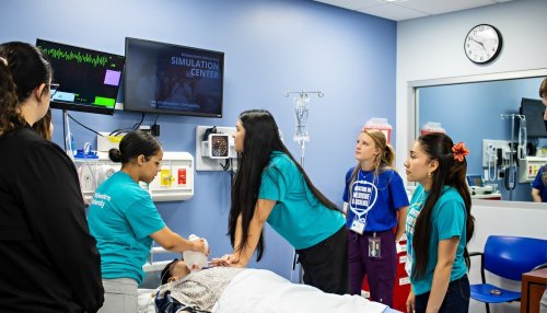 Students in teal MWU shirts practice on a simulation mannequin in the Midwestern University Simulation Center while peers observe.