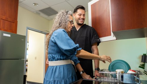 An occupational therapist in black scrubs assists an older patient with daily living tasks in a kitchen therapy setting.